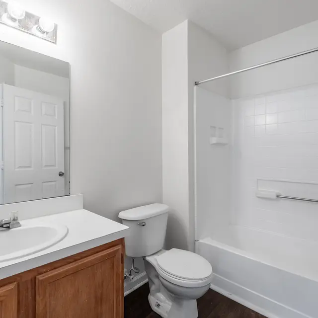 A clean and modern bathroom featuring a white bathtub and shower, a wooden vanity with a sink, a toilet, and a large mirror above the sink. The walls are painted white and the floor appears to be dark wood. There is a door visible in the background leading out of the bathroom.