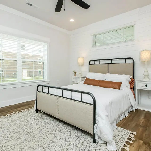 Cozy bedroom with a metal frame bed, white bedding, two nightstands, a window with blinds, and a decorative rug on wooden floor.