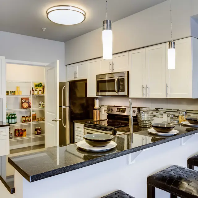 A modern kitchen featuring white cabinets, stainless steel appliances, and a dark granite countertop with bar stools. A pantry can be seen in the background.