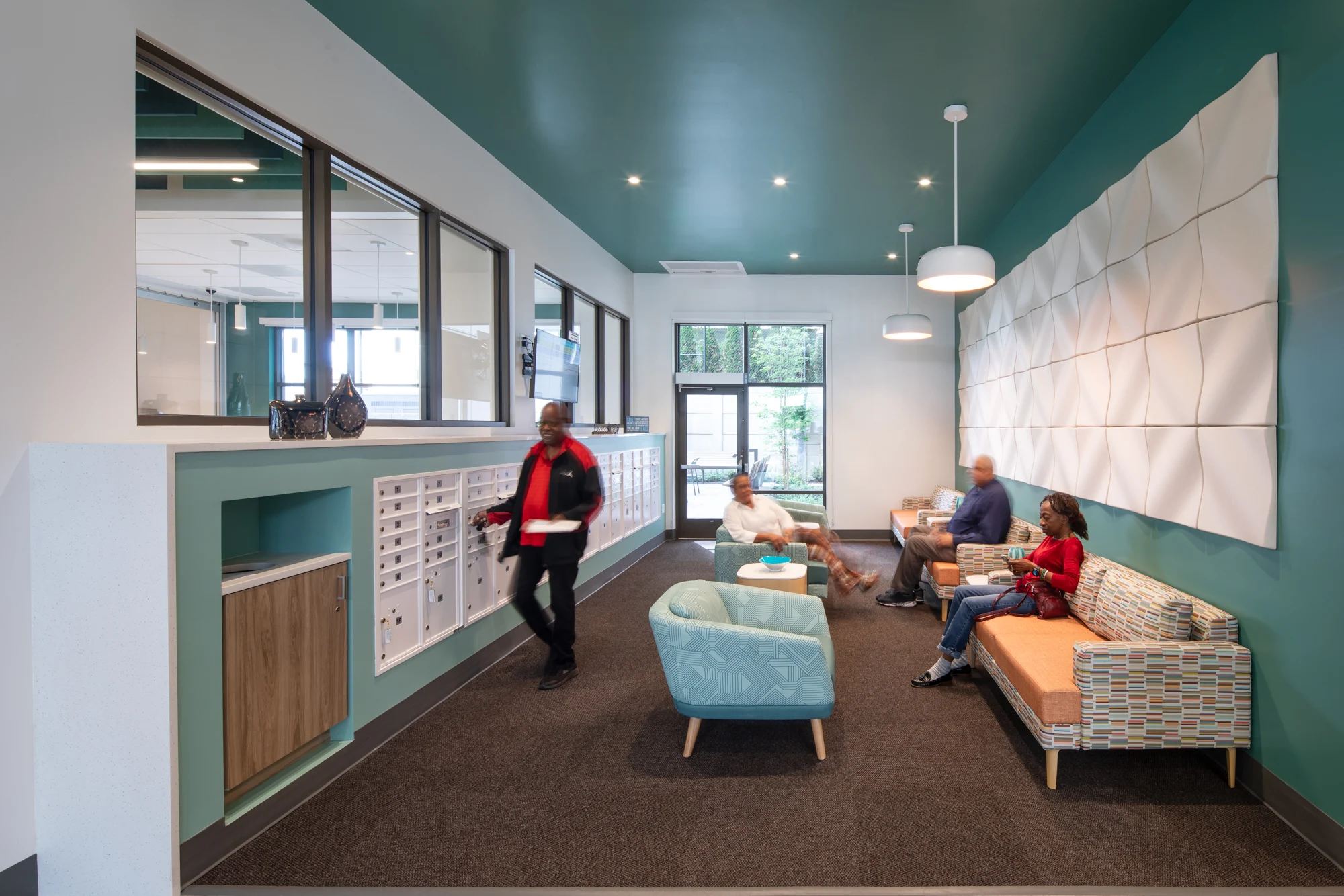 Modern Community Lobby A modern lobby area with turquoise walls, seating arrangements, and a mail station. Several people are seated on couches, and one person is walking in.