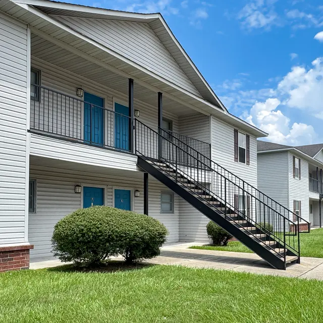 Exterior view of an apartment building with blue doors and black staircases, surrounded by green grass under a partly cloudy sky.