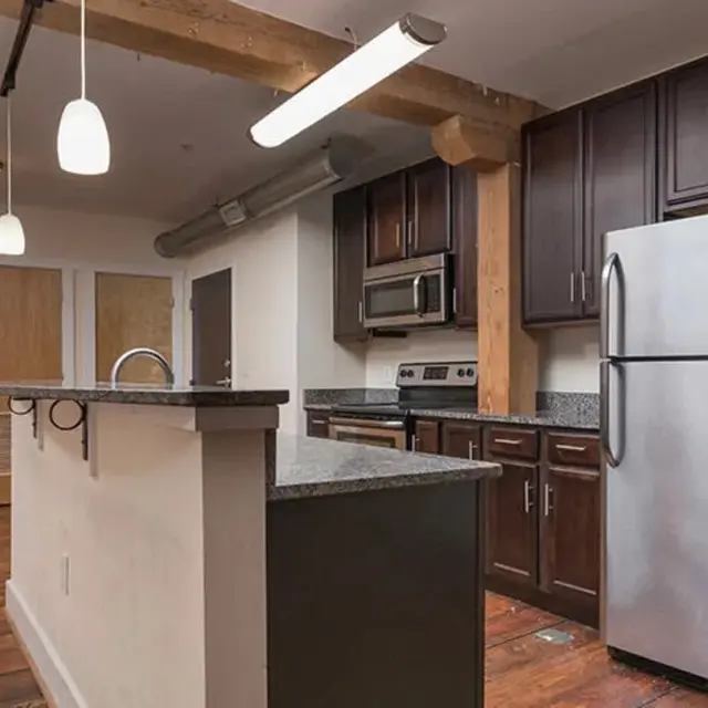 A modern kitchen featuring dark wooden cabinets, stainless steel appliances, and a granite countertop with an island. The space has an open layout with wooden floors and overhead lighting.