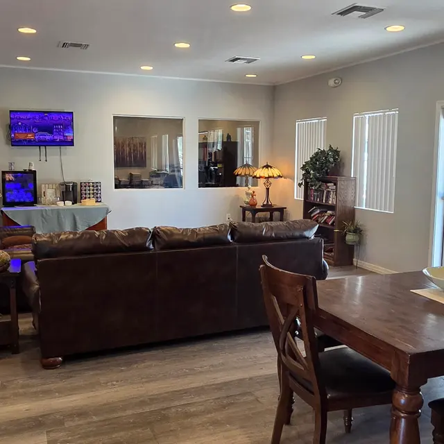 A cozy living room featuring brown leather sofas, a wooden dining table, decorative lamps, and a television mounted on the wall, with natural light coming in through the windows.