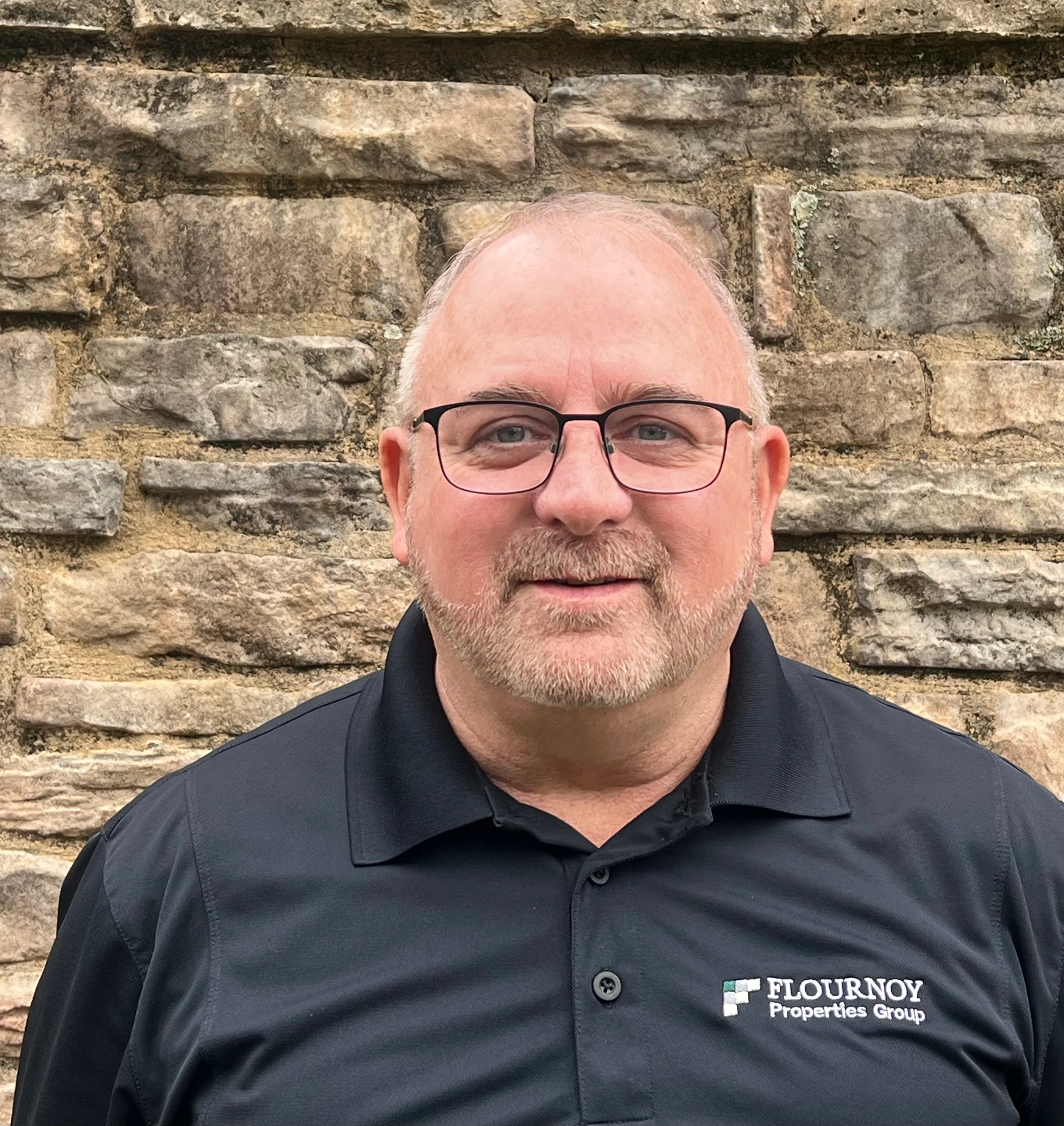 Man Smiling in Front of Stone Wall A man with glasses and short hair is smiling while standing in front of a stone wall. He is wearing a black polo shirt with the logo of Flournoy Properties Group on it.