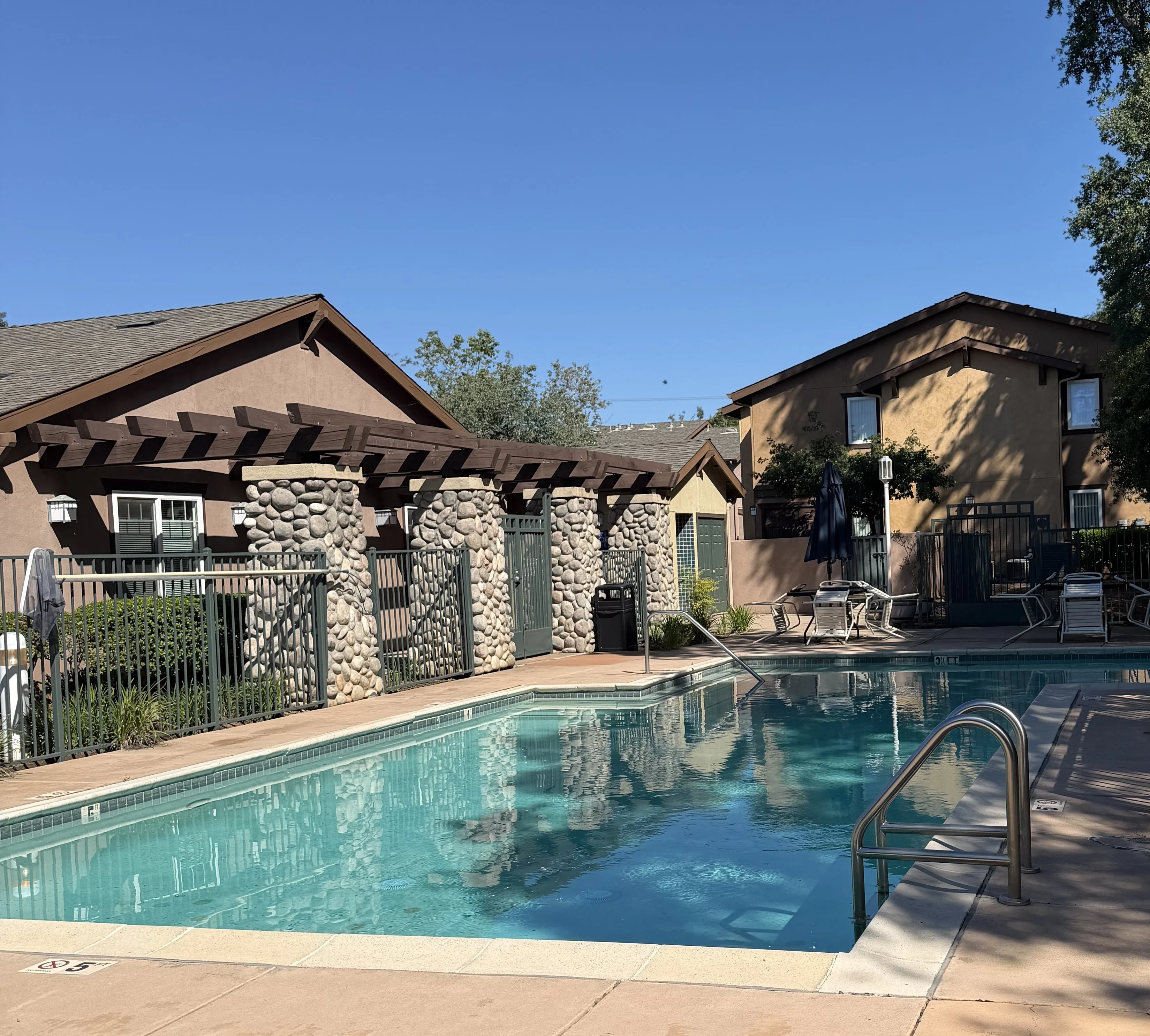 A clear swimming pool with a light-colored deck surrounded by a green lawn and residential buildings. There are lounge chairs beside the pool and a stone archway leading to a gated area.