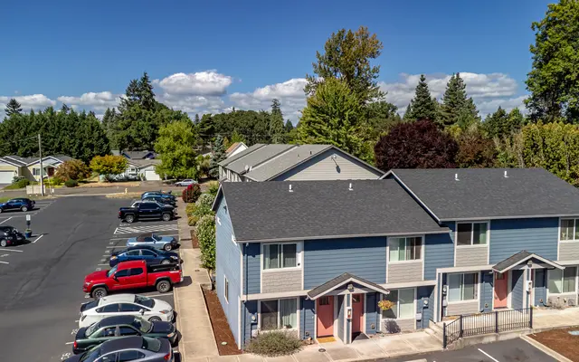 Aerial view of a residential apartment complex featuring two-story blue and grey buildings, surrounded by green trees and a parking area. The scene presents a clear blue sky with some clouds.