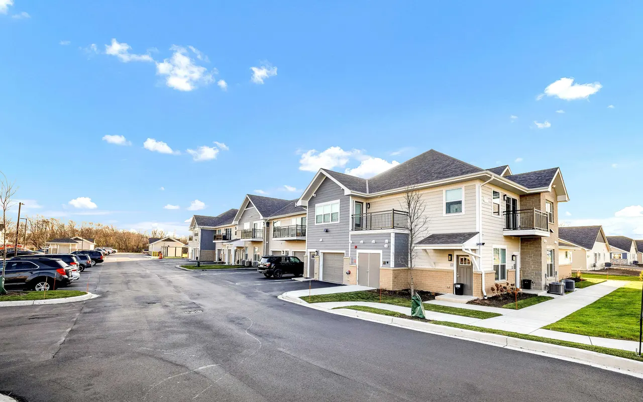 View of a modern apartment complex on a sunny day, with cars parked along the side and green grass areas.