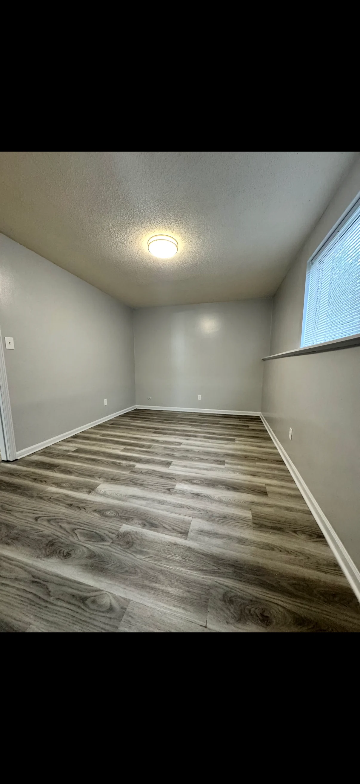 Vacant Room with Natural Light A vacant, unfurnished room with grey walls and wooden flooring, featuring a single window that allows natural light to enter, illuminated by a ceiling light.