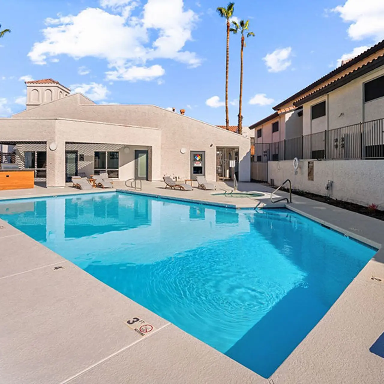 A clear blue swimming pool surrounded by lounge chairs, with a modern building in the background and palm trees nearby.