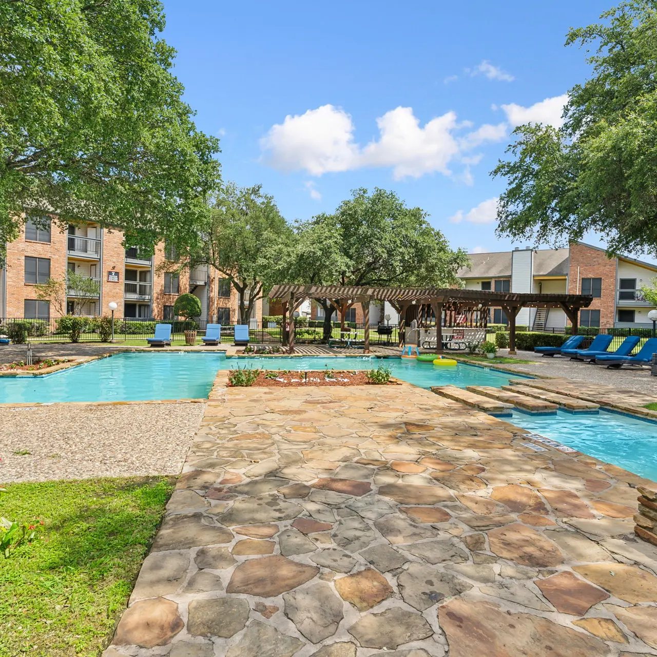A scenic view of a swimming pool area in an apartment complex, featuring lounge chairs, a pergola, and lush greenery.