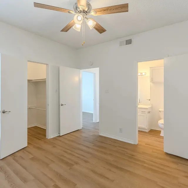 Interior of a modern apartment bedroom with wooden flooring, featuring two open doors leading to closets and a bathroom, along with a ceiling fan and a window with blinds.