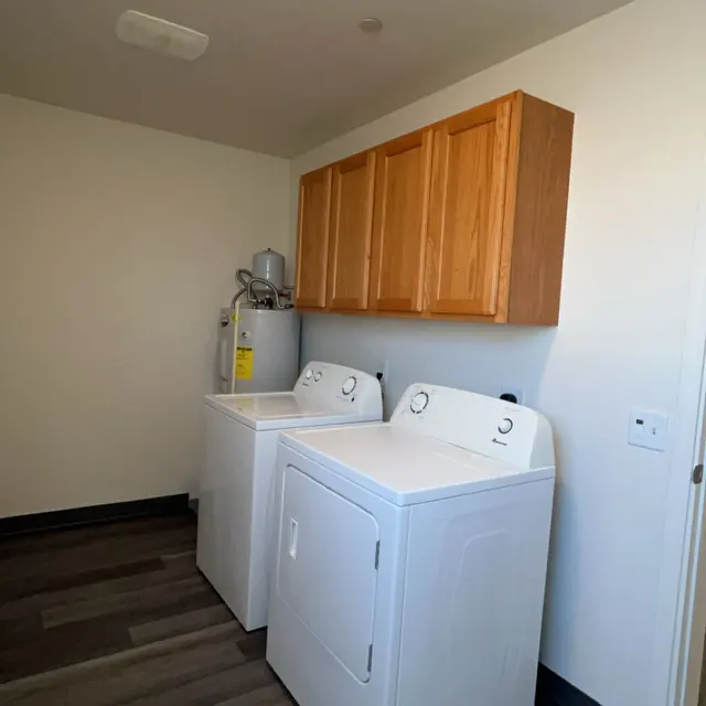 A clean and organized laundry room featuring two white washers and a dryer, along with wooden cabinets above for storage. The floor has a dark wood design, and the walls are painted white.