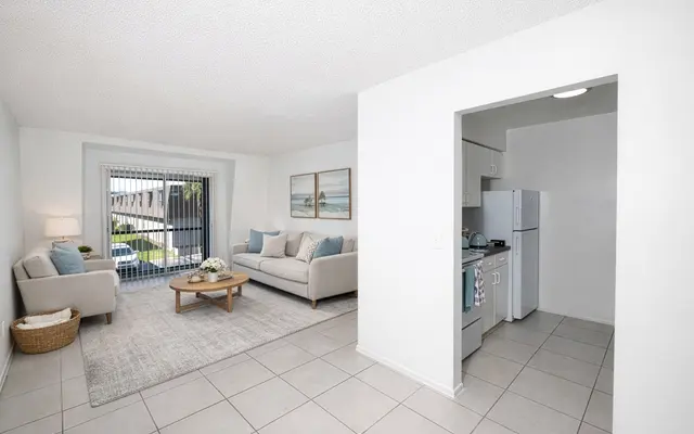 Stylish Living Room and Kitchen A modern living room paired with a kitchen area. The living room features a light-colored sofa and a coffee table on a rug. Large windows allow natural light to fill the room, showcasing a view outside. The kitchen area is visible with light cabinetry and appliances.