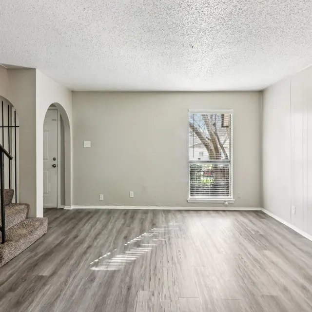 A spacious and empty living room featuring light-colored walls and new flooring. There is a staircase on the left leading to an upper level. A window on the right allows natural light to enter the room.