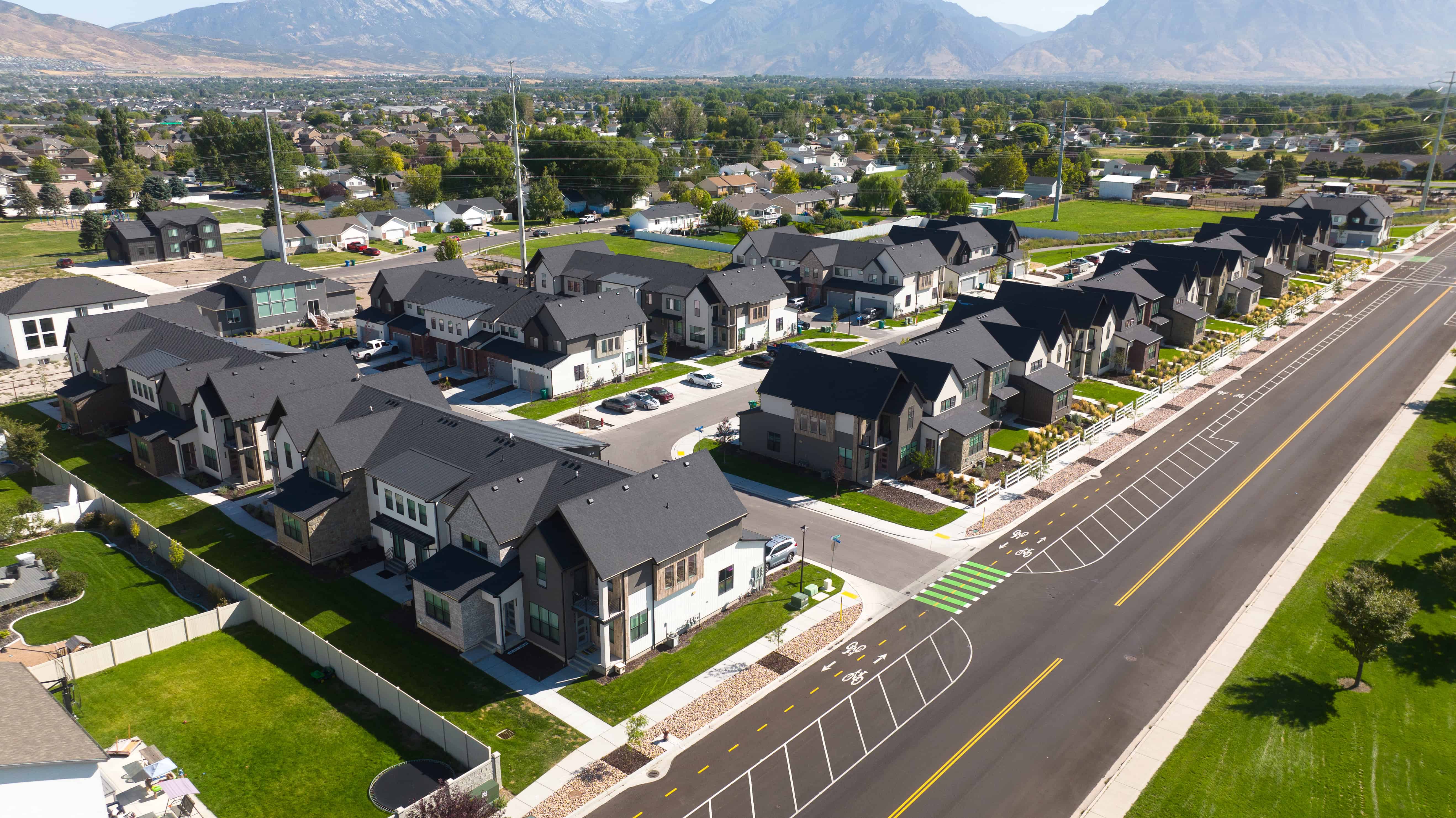 Snow Springs Towns Aerial view of a suburban neighborhood with modern homes, green lawns, and a clear road, surrounded by mountains.