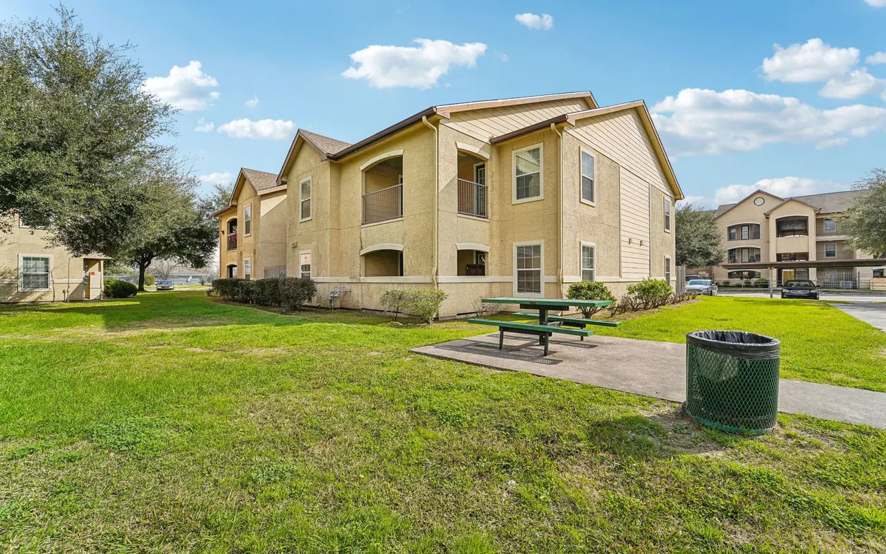 Exterior view of a two-story apartment complex with a yard, featuring picnic tables and a trash can.