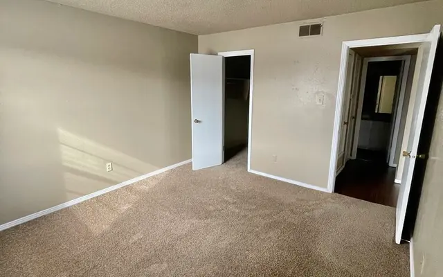 Empty Bedroom Interior A spacious empty bedroom with beige carpet and light-colored walls. There are two doors leading to other areas, with natural light coming in from the left side of the image.