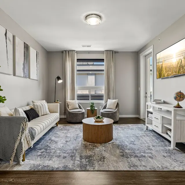 A modern living room featuring a white sofa adorned with pillows, two gray armchairs, a round wooden coffee table, and a decorative plant in a pot. The room is illuminated by natural light through large windows, with artwork on the walls and a television mounted above a white console table.