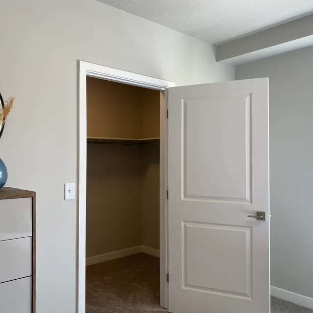A well-lit modern bedroom interior featuring a partially open closet and a decorative dresser with a round mirror and vase
