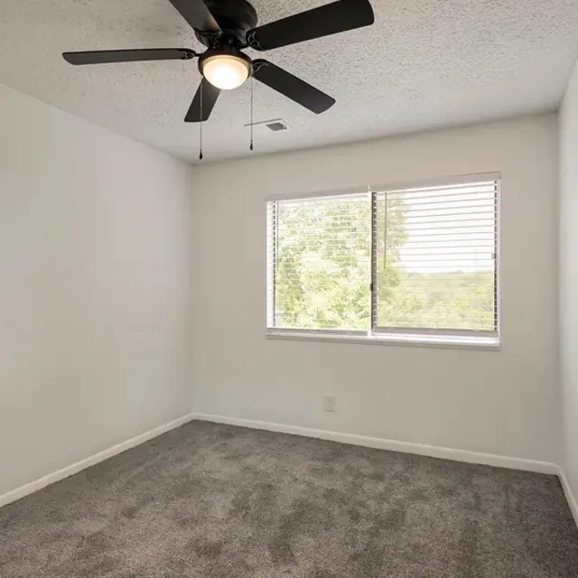 A small, empty bedroom with light grey carpet, a ceiling fan, and a window showcasing a view of trees outside.
