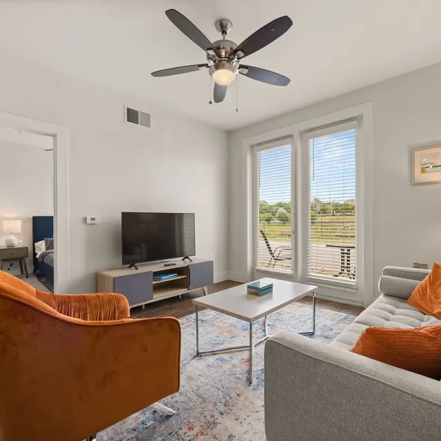 A modern living room with a comfortable couch, armchair, coffee table, and a TV setup near large windows with natural light.