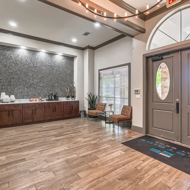 A modern lobby with wooden flooring, a reception area featuring a dark wood counter, decorative wall art, and seating arrangements. There are large entrance doors and large windows allowing natural light in.