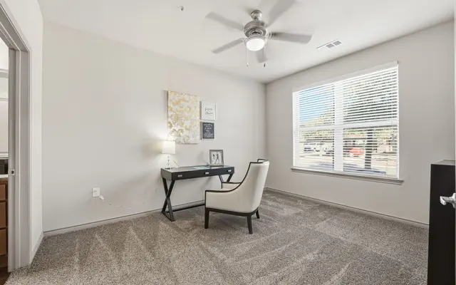 A cozy room with a window, desk, chair, and ceiling fan, featuring neutral walls and carpeted flooring.