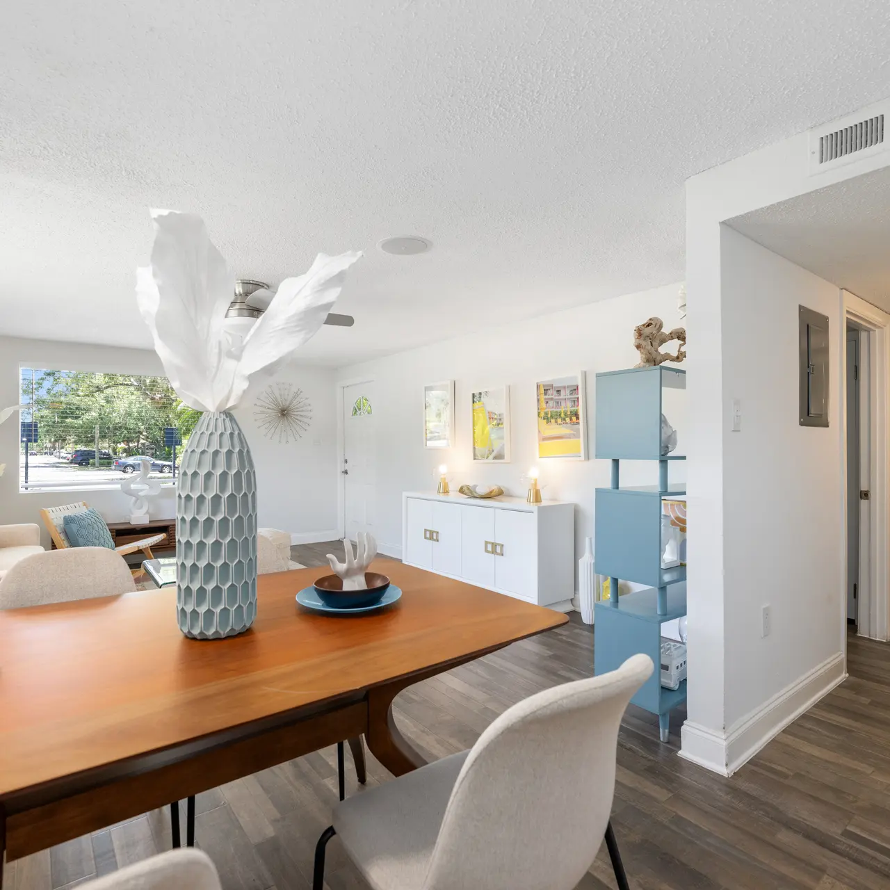 A contemporary living area featuring a wooden dining table with decorative items and modern seating. Natural light flows in through the windows, illuminating stylish decor and a console unit against the wall.
