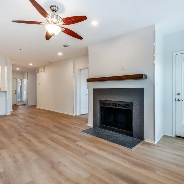 A spacious living room featuring a ceiling fan, hardwood floors, and a fireplace. There is an entry door on the right leading outside, and the decor is minimalistic with light walls.