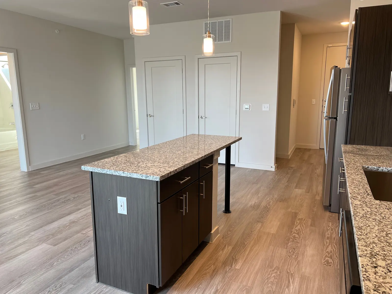 Modern Kitchen Interior A modern kitchen featuring a spacious island with a granite countertop, dark cabinetry, and stainless steel appliances. The area has light-colored walls and wooden floorboards, with bright pendant lighting above the island.
