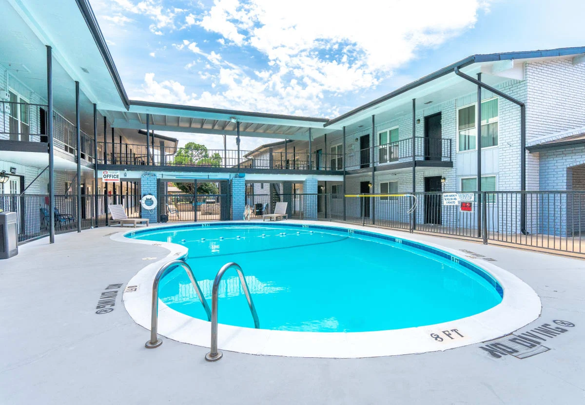 Apartment Complex Pool Area A clear blue swimming pool located in a courtyard surrounded by two-story apartment buildings. The area features a fence around the pool, and there are chairs and tables nearby for relaxation.