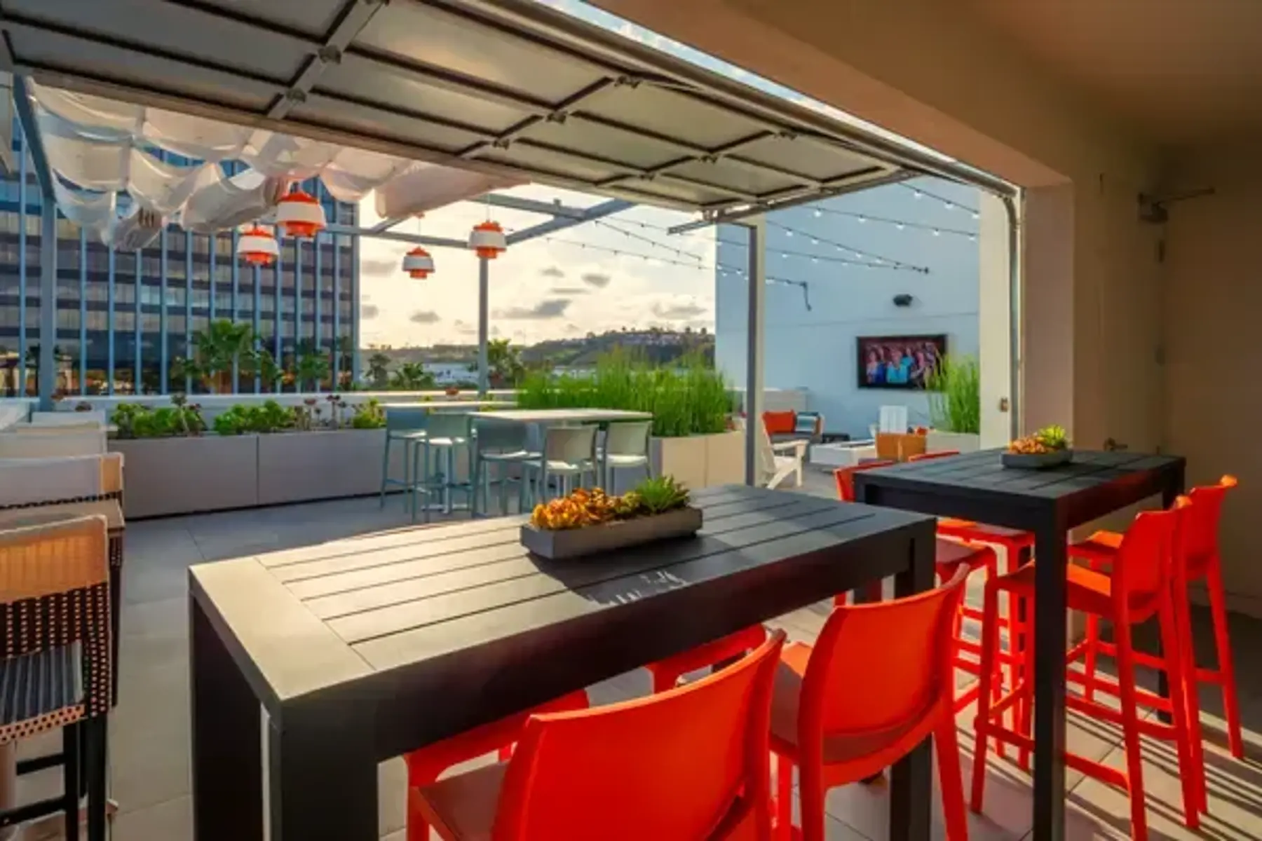 Stylish Rooftop Lounge A modern rooftop lounge with bright orange chairs and a black table. The area features a green plant arrangement, string lights overhead, and city buildings in the background. There is a TV on the wall and additional seating in the distance.