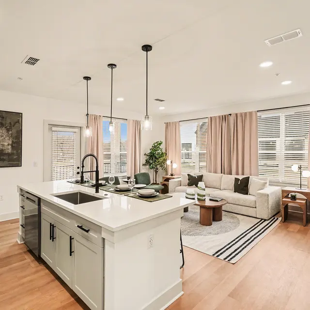 A modern living room and kitchen area featuring a light-colored kitchen island with a sink, pendant lights, a cozy seating area with a sofa and coffee table, and large windows with curtains.