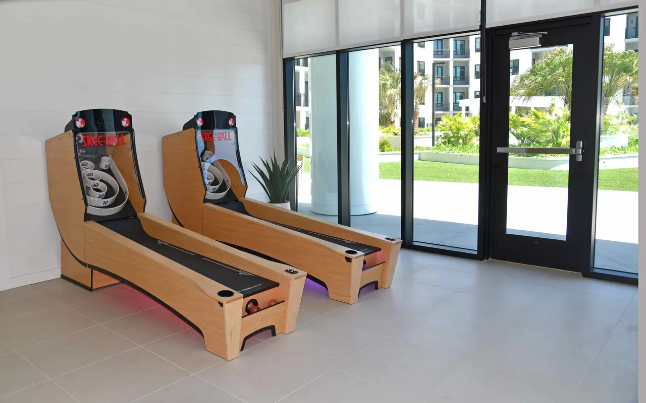 Two skee ball arcade machines in a brightly lit indoor area, with a glass door leading outside and a view of greenery.