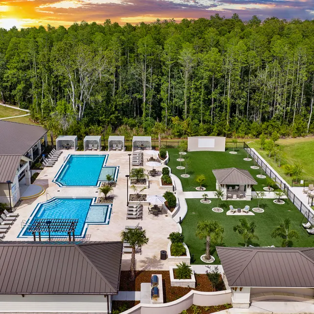 Aerial View of Luxury Pools and Green Spaces Aerial view of a residential area featuring two swimming pools surrounded by lounge chairs and cabanas, alongside a well-maintained green space, landscaped areas, and a scenic background of trees at sunset.