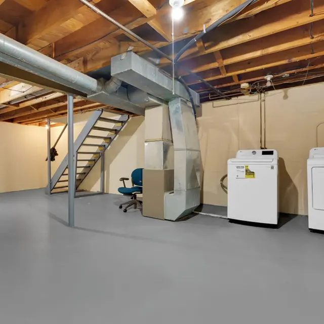 A spacious basement with bare walls, wooden beams, and a concrete floor. There's a staircase leading up, a green chair, a cardboard box, a washing machine, and a dryer. The space is illuminated by overhead lights.
