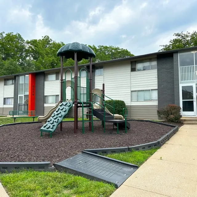 A playground with a slide and climbing structure in front of an apartment complex, surrounded by mulch, with green grass and trees in the background.