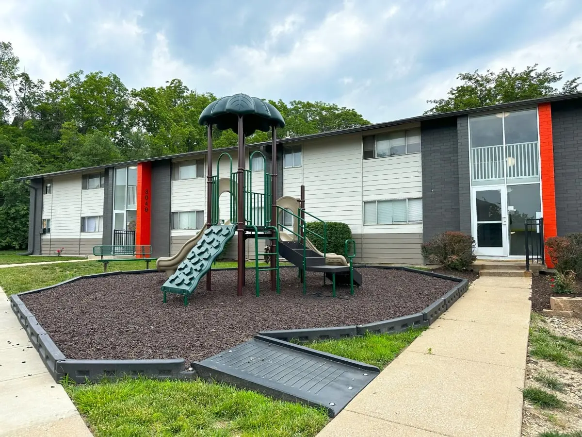 A playground with a slide and climbing structure in front of an apartment complex, surrounded by mulch, with green grass and trees in the background.