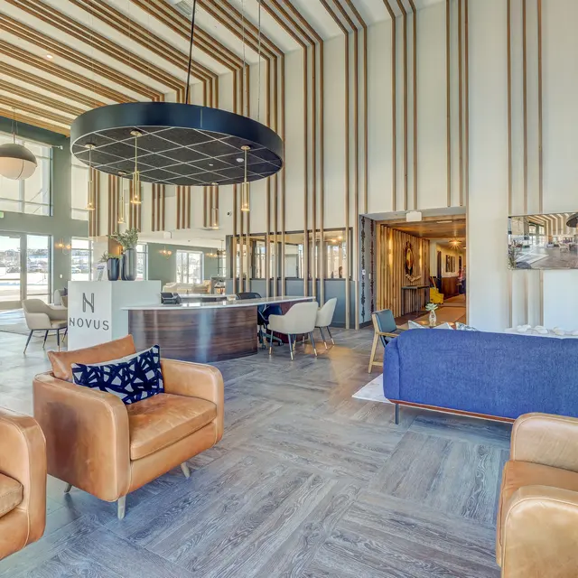 A spacious and modern lobby featuring brown leather seating, a large round chandelier, and wooden wall accents. The lobby has large windows, allowing natural light to flood the space.