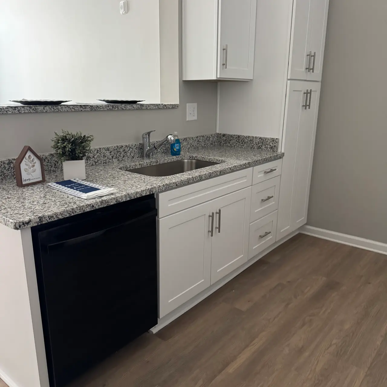 A modern kitchen featuring white cabinets, a granite countertop, and a black dishwasher. The kitchen has a sink with a faucet and a decorative plant on the counter.