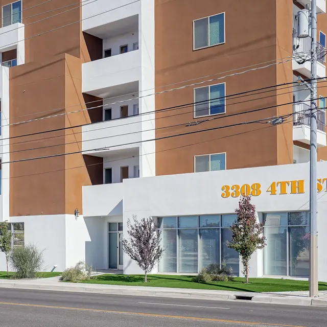Contemporary Apartment Building A modern multi-story apartment building with a white and brown facade. The ground floor features large glass windows and a bold address sign. There are green lawns and trees in front, and utility poles with wires visible in the background.