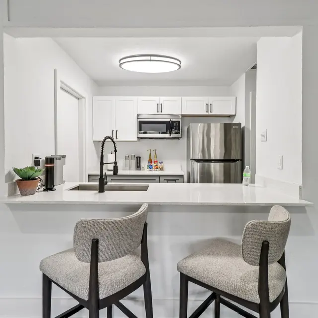 A modern kitchen with a white color scheme, featuring a counter bar with two high chairs, stainless steel appliances, and contemporary lighting.