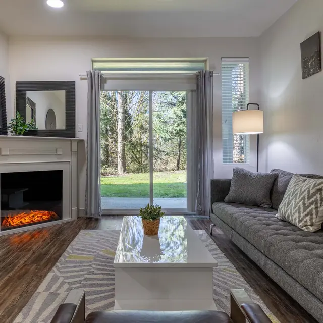 A cozy modern living room featuring a gray sofa with decorative pillows, a white coffee table, and a fireplace. Large windows offer a view of greenery outside. The room is illuminated with natural light and has a stylish lamp.