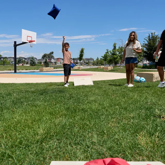 Playtime at the Park A child throws a blue pillow while two adults stand nearby in a park with a basketball hoop in the background and a clear blue sky.