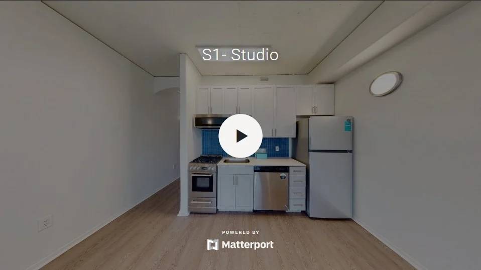 A studio apartment featuring a small kitchen area with an oven, refrigerator, and sink, against a simple wall with light-colored flooring.