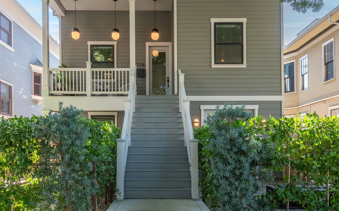 A two-story gray house with a porch, flanked by green hedges and trees, on a sunny day.