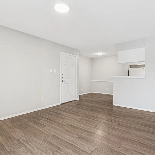 An empty apartment living room with light hardwood flooring, beige walls, and a white door. There is a small opening that leads to a kitchen area in the background, with gray countertops and a breakfast bar.