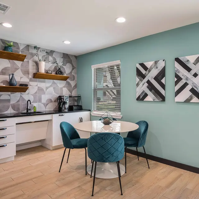 A modern kitchen and dining area featuring a light blue wall, patterned wallpaper, a round white dining table with teal chairs, and shelves with decorative items.