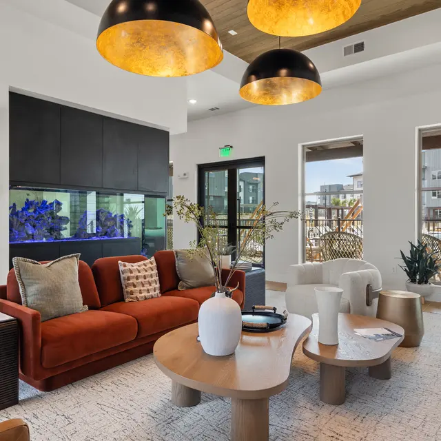 A stylish and modern living room featuring a cozy red sofa, a variety of seating options, and large windows letting in natural light. There is an aquarium and decorative plants, along with contemporary light fixtures. The decor is chic and inviting.