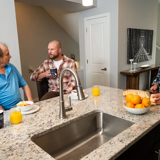 Morning Discussion in a Modern Kitchen A casual kitchen scene with three people sitting around a countertop. Two men are having a discussion while a woman observes, all enjoying breakfast items and drinks.